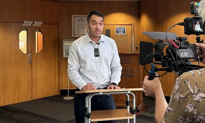 A man in a light shirt speaks at a podium indoors, facing a video camera and microphone, with wooden doors and signs in the background. Another person is partially visible operating the camera.