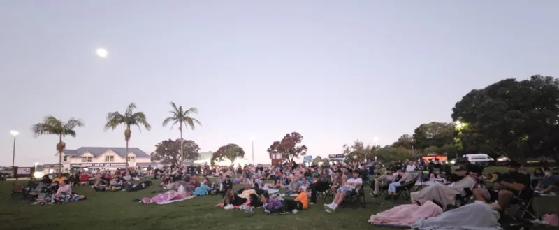 A photo of a twilight scene: people are nestled on picnic blankets on grass with palm trees in the background.
