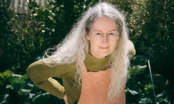 A photo of the writer Elizabeth Knox who has long grey-white hair, is wearing salmon coloured dungarees and is looking up at the camera from where she's standing outside in front of a garden. It is sunny.