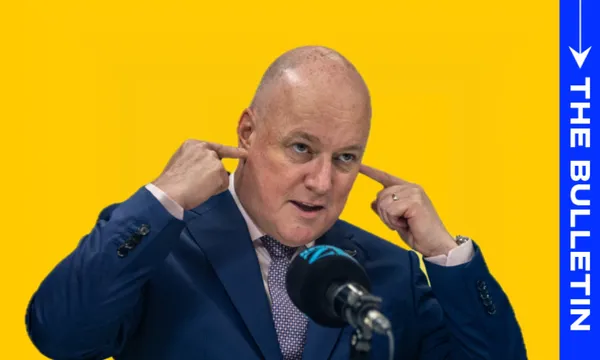 A bald man in a blue suit gestures by plugging his ears with his fingers while speaking into a microphone. The background is bright yellow, with a vertical blue banner reading "The Bulletin" on the right.