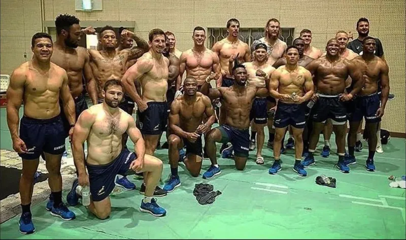 A group of muscular men in navy blue athletic shorts and shoes pose together indoors, many smiling and some flexing. They stand and kneel on a green mat in a brightly lit room.