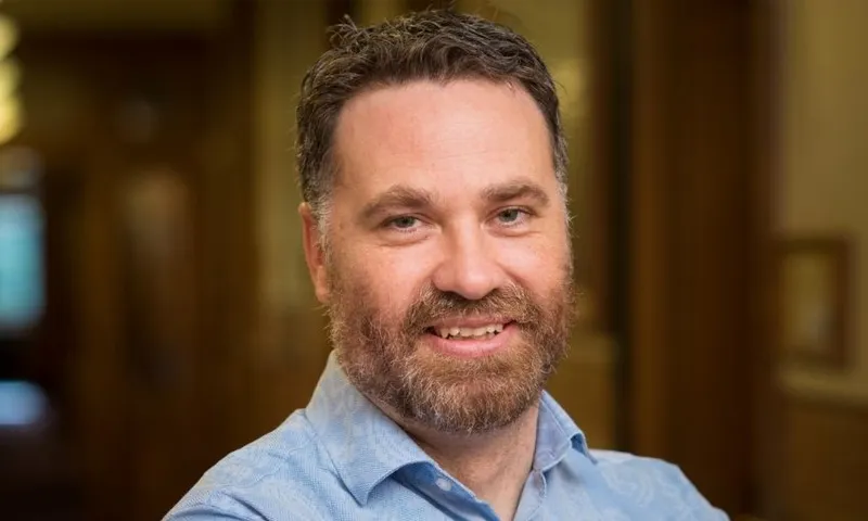 A man with short curly brown hair and a beard smiles at the camera. He is wearing a light blue collared shirt and is standing indoors with a softly blurred background of wooden walls and warm lighting.