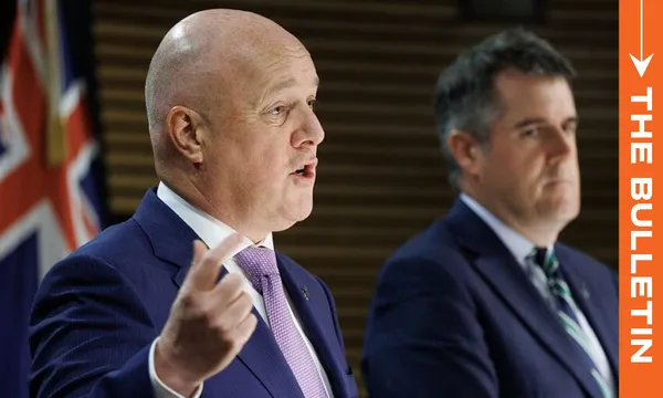 Two men in suits stand in front of a New Zealand flag. The man in the foreground is speaking and gesturing with his hand. An orange banner on the right reads "THE BULLETIN.