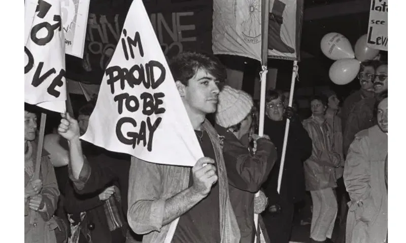A black and white photo of a protest where the protest sign says "proud to be gay"