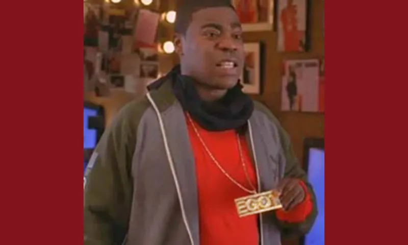 A TV still from 30 Rock, showing Tracy Jordan wearing a red t-shirt with black headphones around his neck. He's holding a heavy gold chain with a pendant and EGOT on it