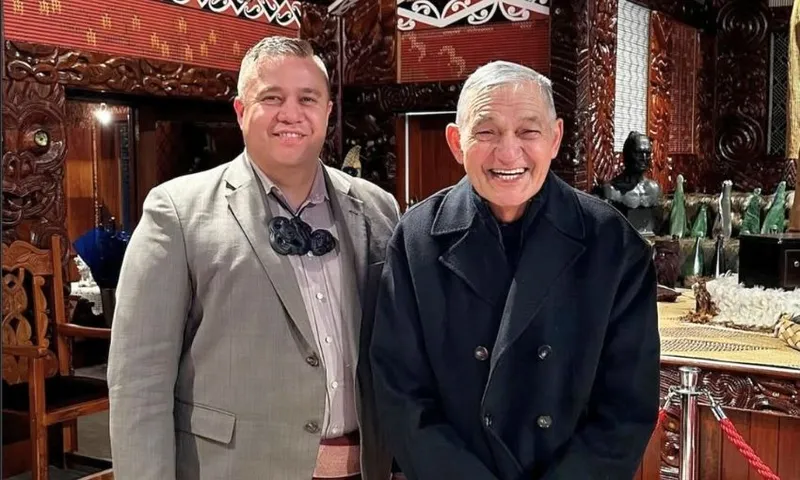 Two men stand smiling indoors, surrounded by ornate carved wooden decorations and traditional Māori art. One wears a grey suit with a necklace; the other wears a dark coat. The setting appears formal and cultural.