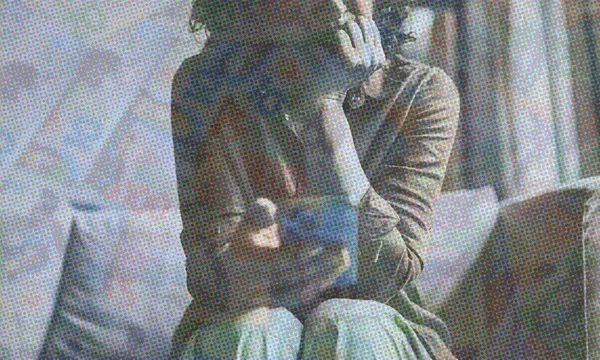 A person sits on a couch, leaning forward with their chin resting on one hand, holding a smartphone in the other. The image has a grainy, textured effect and muted colors.