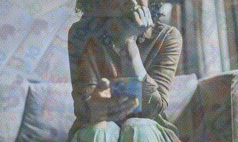 A person sits on a couch, leaning forward with their chin resting on one hand, holding a smartphone in the other. The image has a grainy, textured effect and muted colors.