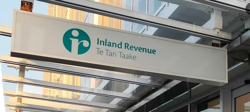 A sign for Inland Revenue, Te Tari Taake, hangs under a glass canopy outside a building. The sign features a teal logo and text.