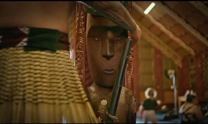A person in traditional Māori attire stands beside a carved wooden figure inside a meeting house, holding a black staff. The background shows others practicing or interacting in the space.