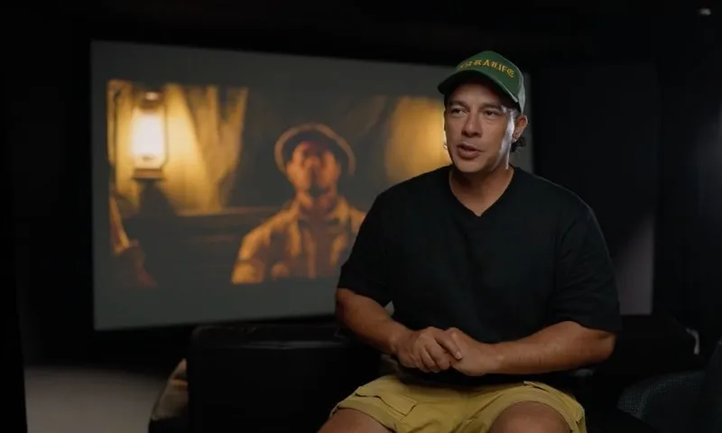 A man in a dark room sits and talks, wearing a green cap, black t-shirt, and yellow shorts. Behind him is a large screen showing a movie scene with a person illuminated by a lantern.