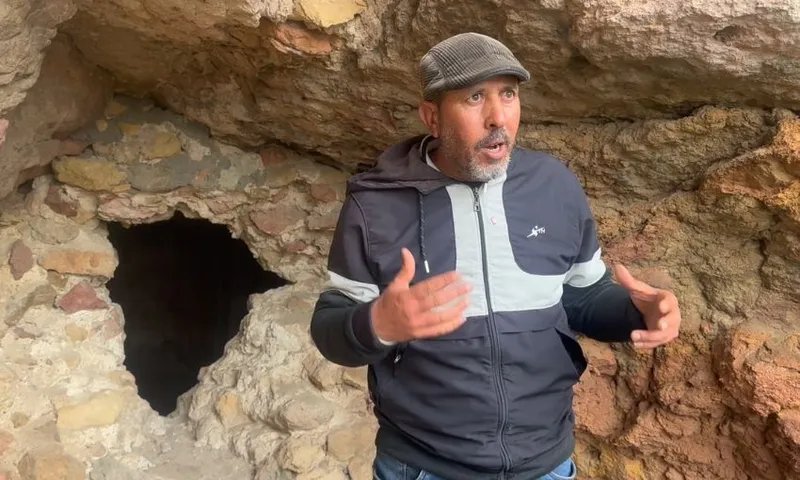 A man wearing a gray flat cap and black and gray jacket gestures with his hands while standing in front of a rocky wall with a dark cave opening.