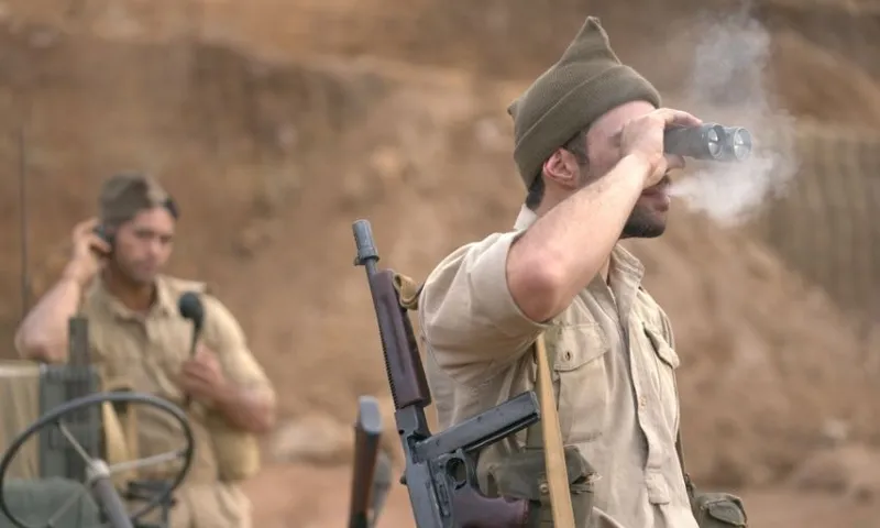 A soldier in WWII-style uniform uses binoculars while exhaling visible breath, with another soldier behind him using a field radio. Both are outdoors in a barren, earthy landscape.