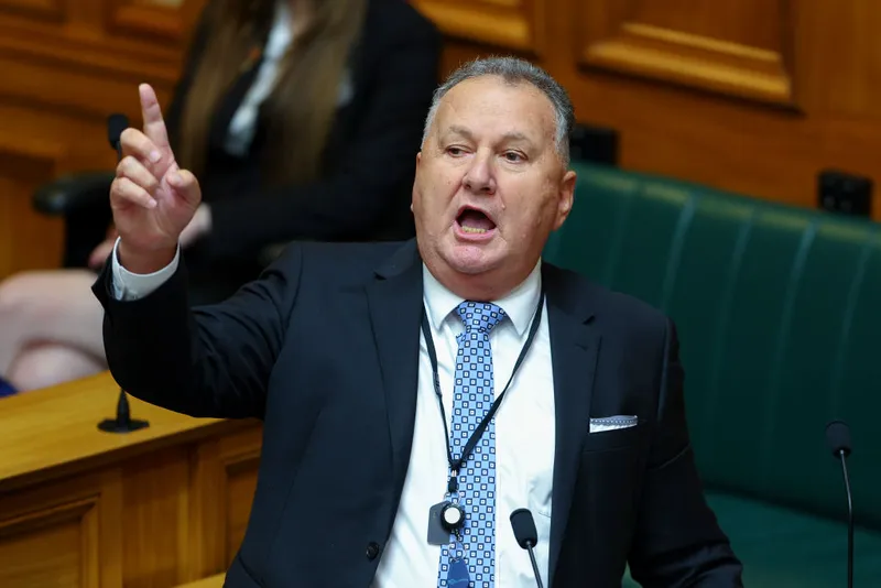 A man in a suit speaks passionately while raising his right hand and pointing upward in a government or parliamentary setting.