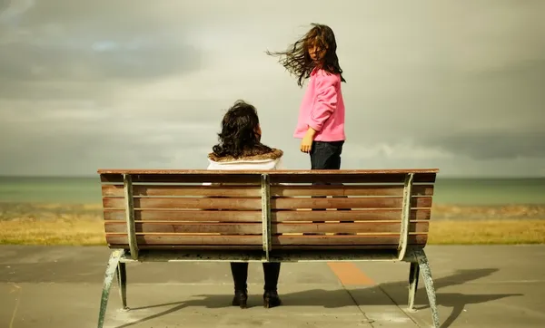 A woman sits on a wooden bench facing the ocean, while a girl in a pink sweater stands on the bench, her hair blowing in the wind. The sky is cloudy and the scene feels calm and contemplative.