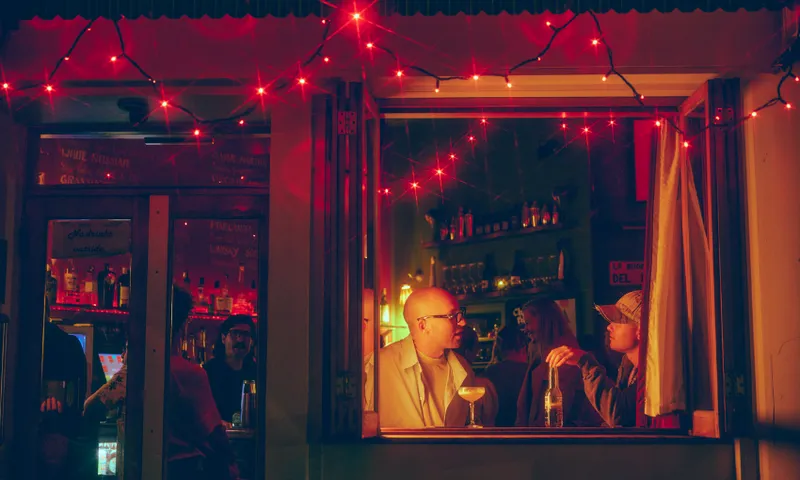 Two people sit by an open window at a dimly lit bar, talking over drinks. Warm yellow light illuminates them, while red string lights hang above, creating a cozy, intimate evening atmosphere.