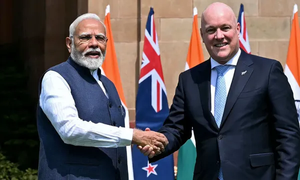 Two men in formal attire shake hands outdoors, standing in front of Indian and New Zealand flags.