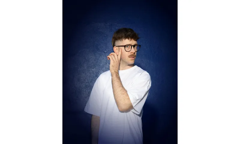 A photo of a young man with glasses and a moustache, wearing a white t-shirt on a dark navy background.