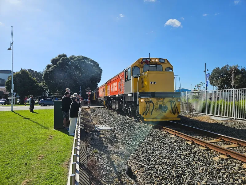 A yellow and orange locomotive approaches a crossing as people stand nearby on a sunny day, with blue sky and a grassy area to the left.