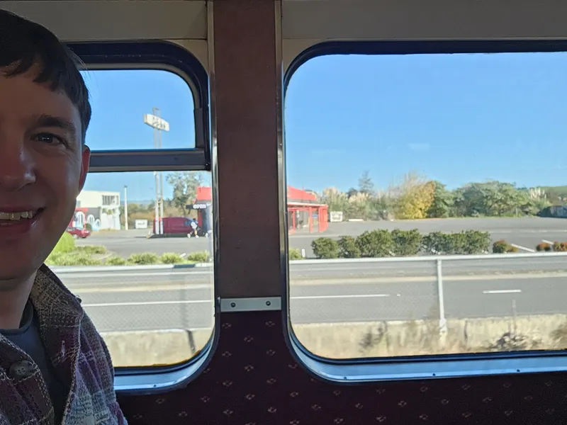 A man smiling sits on a train next to a window; outside, there’s a clear blue sky, a red building, some greenery, and an empty parking lot across a road.
