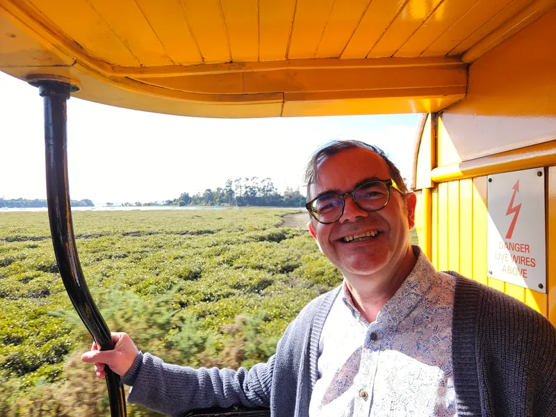 A man with glasses and a gray sweater smiles while riding a yellow tram. Green fields and trees stretch out in the background under a bright, sunny sky. A sign warning of live wires is visible on the tram.