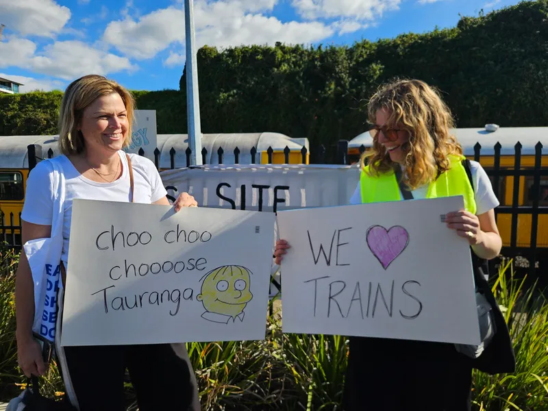 Two women stand outdoors smiling and holding handmade signs. One sign reads "choo choo choooose Tauranga" with a cartoon drawing, and the other reads "WE ♥ TRAINS." There is a train and greenery in the background.