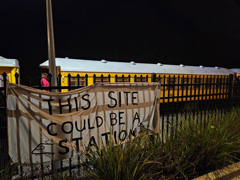 A yellow train stands behind a black metal fence at night. A white cloth banner on the fence reads, "THIS SITE COULD BE A STATION" in large black letters. Some plants grow in front of the fence.