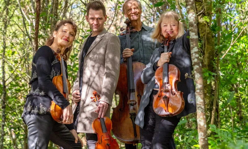 A photo of four classical musicians each holding their instruments and standing in the bush.
