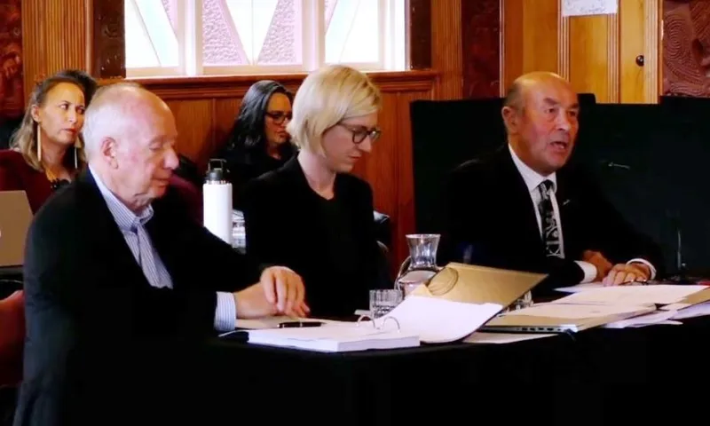 Three people in formal attire sit at a table with documents and microphones during a meeting or hearing, while others sit in the background in a wood-paneled room with decorative windows.