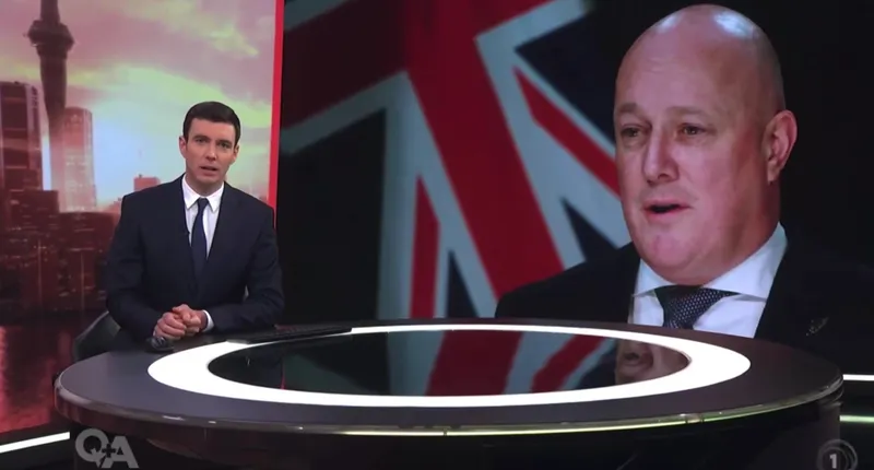 A news anchor in a suit sits at a round desk with city buildings in the background. Beside him is a large screen showing a man in front of a partial New Zealand flag.