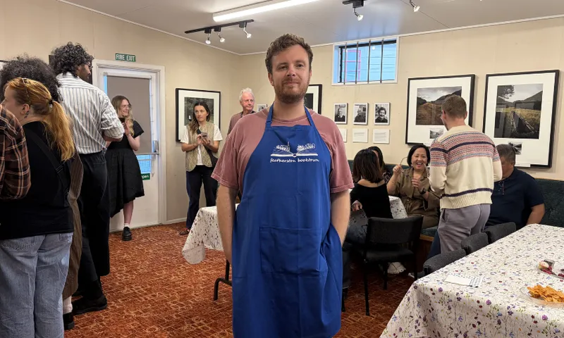 A photo of Jordan Hamel who is a young man wearing a bright blue apron standing in a room full of people talking.