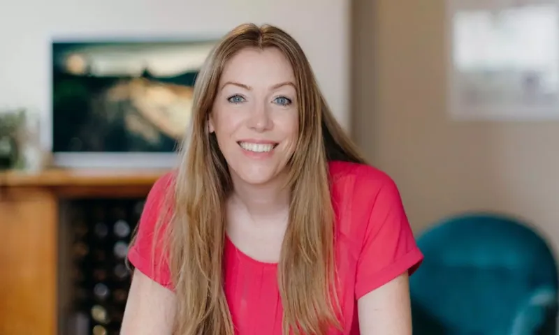 A woman with fair skin, blue eyes and long strawberry blonde hair smiles at the camera wearing a pink short sleeved shirt
