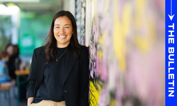 A woman with long dark hair, wearing a black blazer and tan pants, smiles while leaning against a colorful wall. The words "THE BULLETIN" appear vertically on a blue strip on the right side of the image.