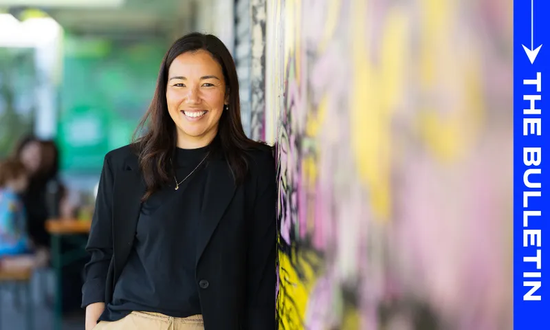 A woman with long dark hair, wearing a black blazer and tan pants, smiles while leaning against a colorful wall. The words "THE BULLETIN" appear vertically on a blue strip on the right side of the image.
