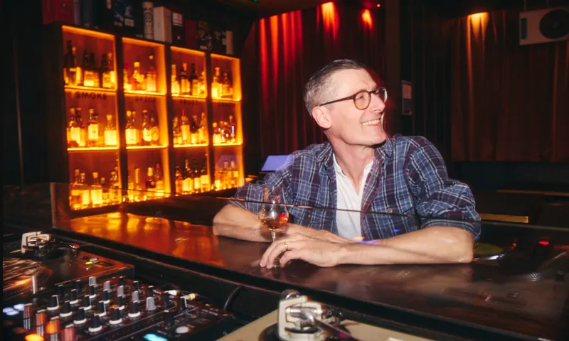 A man wearing glasses and a plaid shirt sits at a bar holding a drink, smiling. Behind him are shelves filled with bottles, softly lit by warm yellow lights. DJ equipment is visible in the foreground.
