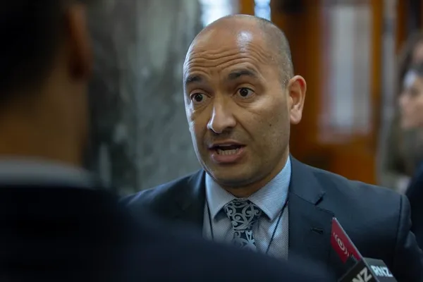 A man in a suit speaks while being interviewed by reporters indoors. He appears engaged and expressive, with microphones visible in the foreground.