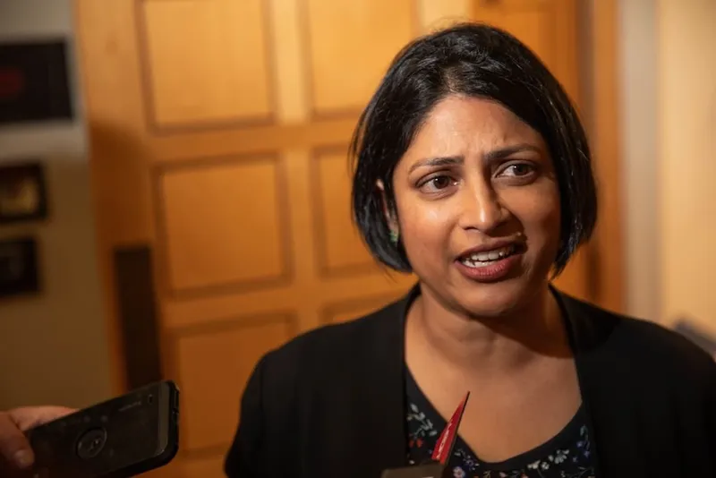 A woman with short dark hair speaks to reporters, holding microphones and recording devices up to her. She is indoors, standing in front of a wooden door, and appears to be responding to questions.