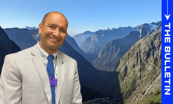 A man in a light gray suit and purple tie smiles in front of a scenic mountain valley under a blue sky. On the right, a vertical blue banner reads "THE BULLETIN" in white letters.