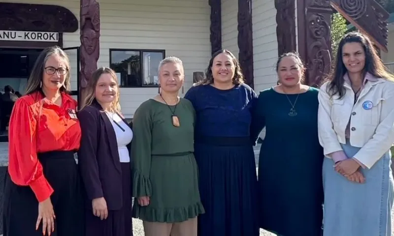 Six women stand side by side, smiling in front of a traditional building with carved wooden pillars and a sign reading "NGA WAHINE O TE MANU-KOROKI." They are dressed in a variety of colorful outfits.