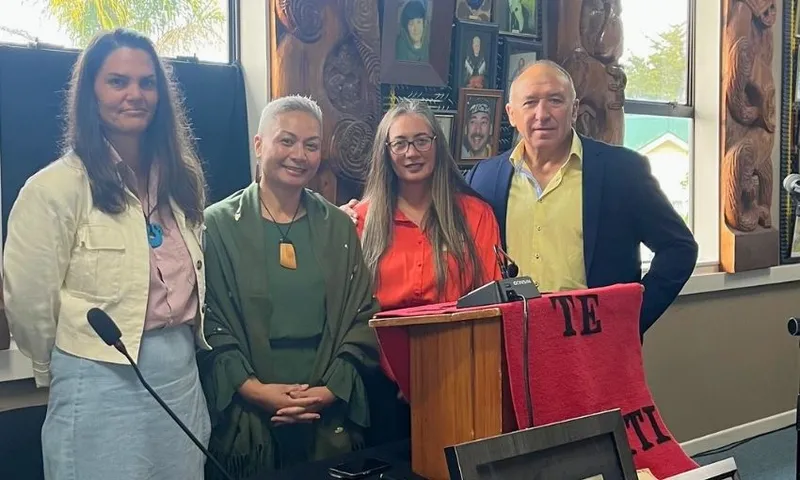Four people stand indoors beside a wooden podium draped with a red cloth reading "TE." Behind them are carved wooden panels and framed photos on the wall. They are smiling, appearing at an event or ceremony.