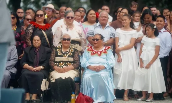 A large group of people, including women in formal dresses and traditional attire, pose together outdoors. Some are seated while others stand behind them, smiling under sunny weather.