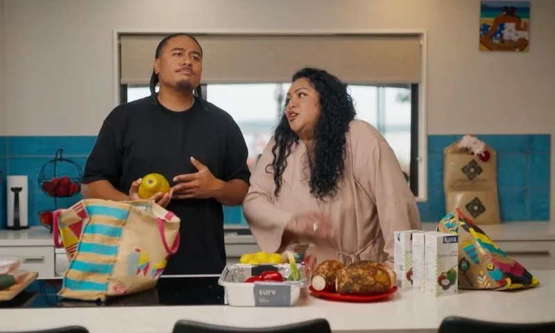 A man and woman stand in a modern kitchen. The man holds an apple while the woman gestures animatedly. Groceries, including fruit and cartons, are spread on the counter in front of them.
