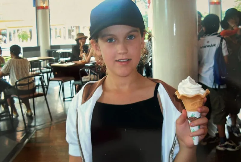 A young girl stands in a shopping mall holding a soft serve cone and wearing a dark cap and white sweater