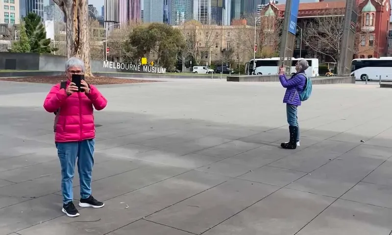 Two older women with white hair stand 10 metres apart and facing different directions. They are each wearing sneakers, jeans and a puffer jacket – one purple and the other pink
