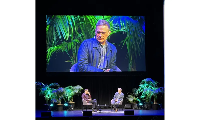 Two men on stage with a large screen behind them showing one of the men close up.