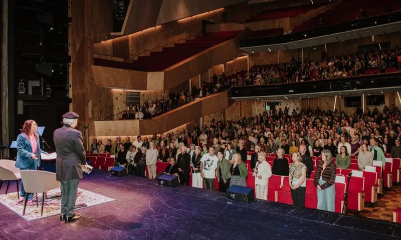 Two people standing on a large stage with a large audience all standing.