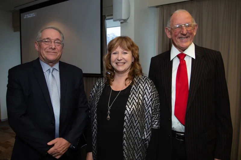 Three adults stand side by side indoors, dressed in formal attire. Two men wear suits and ties, and a woman in the middle wears a patterned blazer and necklace. They are smiling, with a projection screen in the background.