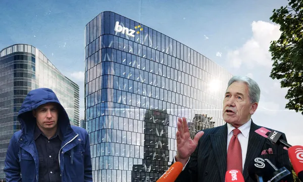 A man in a blue hooded jacket stands near a business-suited man speaking at microphones, with a modern glass building labeled "bnz" in the background under a sunny sky.