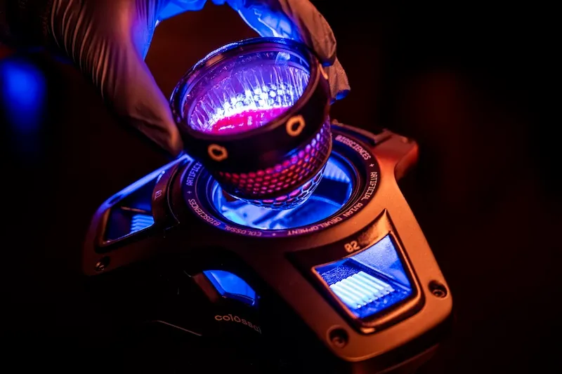 A gloved hand holds a cylindrical object with red lights over a futuristic device emitting blue light, creating a high-tech, laboratory-like atmosphere.