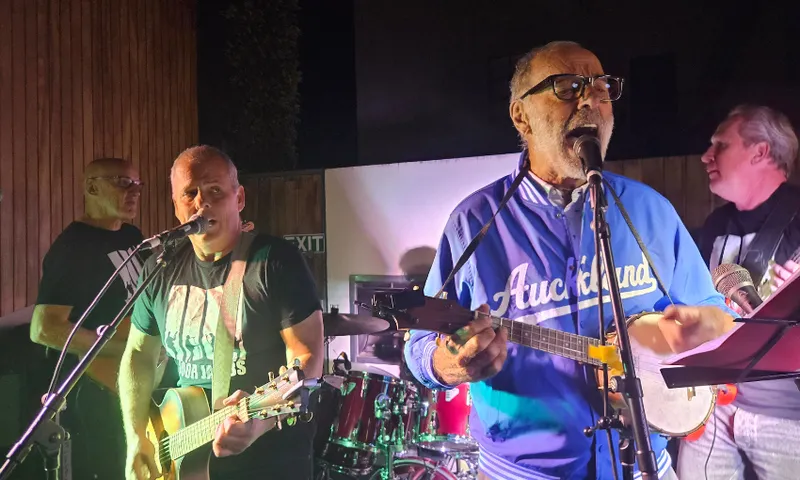 Four older men perform music on stage at night. Three play guitars while singing into microphones, and one plays drums in the background. The man in front wears glasses and a blue "Auckland" jacket.
